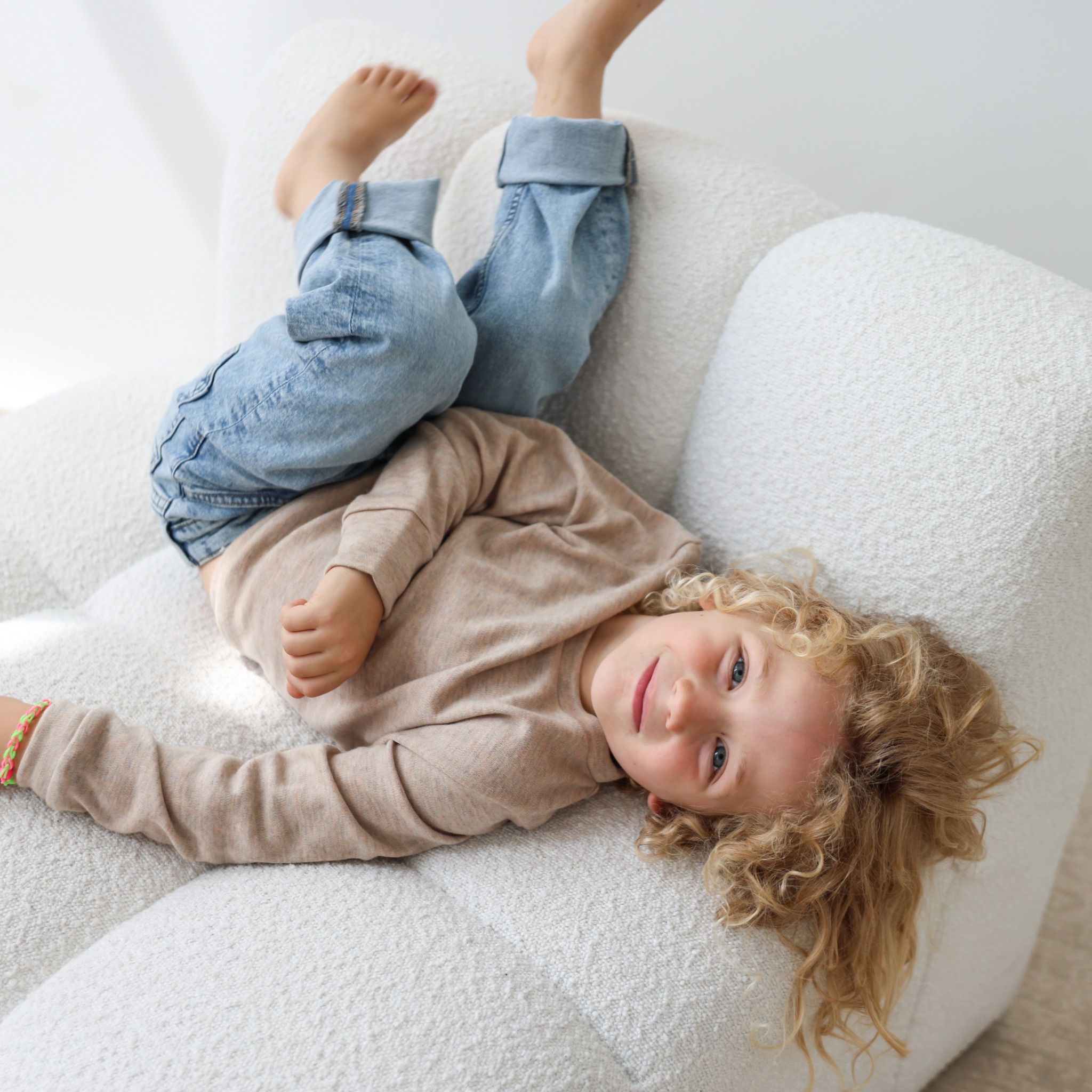 Uśmiechnięty chłopiec z kręconymi włosami leży do góry nogami na białym, teksturowanym fotelu, ubrany w dżinsy i beżową bluzkę. Jasne, naturalne światło.