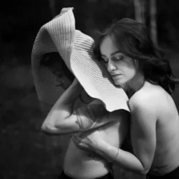 Czarno-białe, artystyczne ujęcie dwóch kobiet w dużym kapeluszu, z subtelnymi tatuażami, w naturalnym oświetleniu, emanujące intymnością i spokojem.