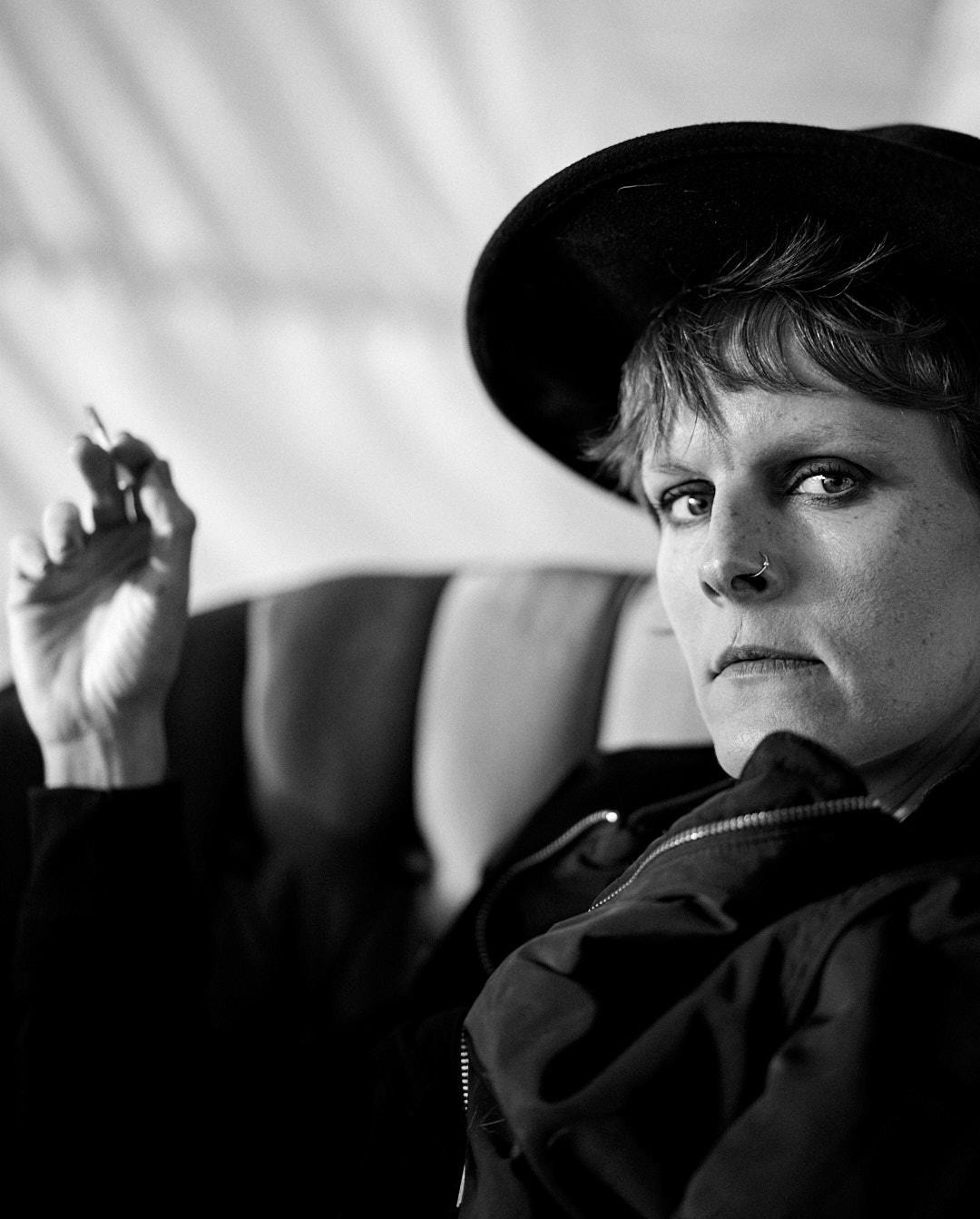 Czarno-biały portret kobiety w kapeluszu, z papierosem w dłoni, siedzącej na fotelu. Wyraziste spojrzenie, piercing w nosie, kurtka z zamkiem błyskawicznym.