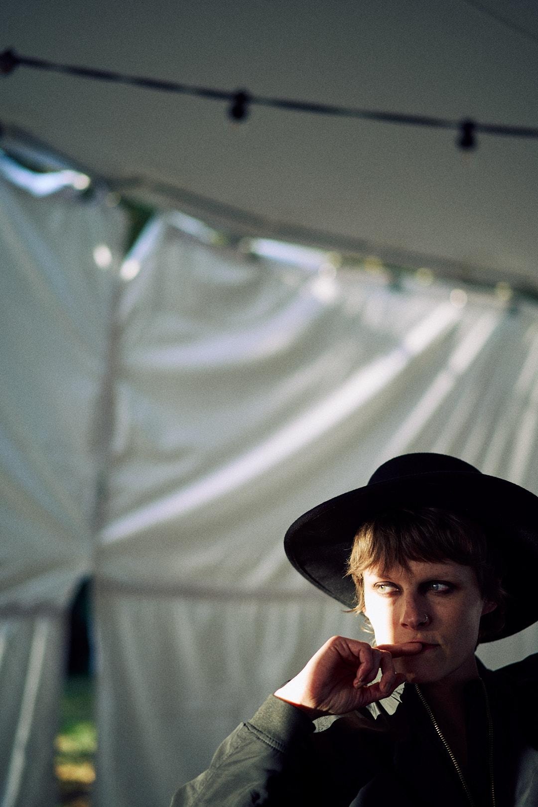 Portret kobiety w czarnym kapeluszu, zamyślona poza, oświetlona naturalnym światłem, na tle białego materiału i delikatnych światełek.