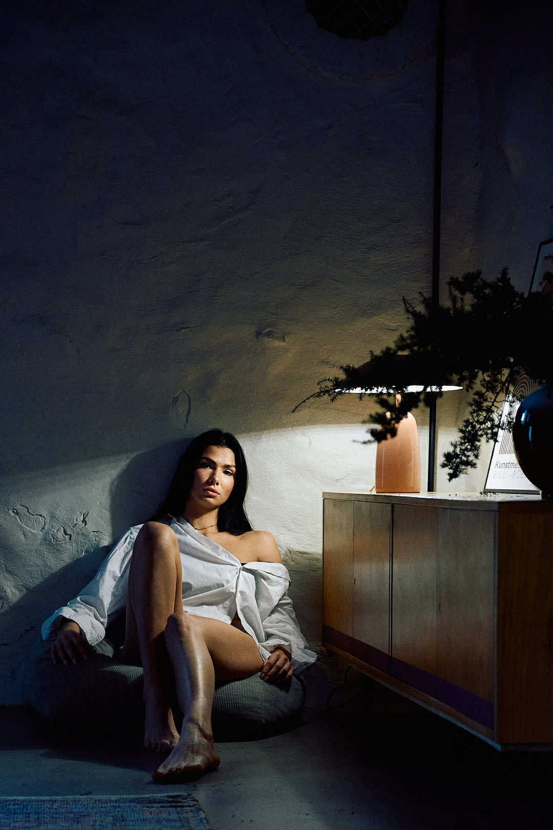 Kobieta w rozpiętej białej koszuli pozuje w ciemnym pokoju z lampą i komodą. Stonowana kolorystyka, surowa ściana, intymna atmosfera. Sesja w minimalistycznym stylu.