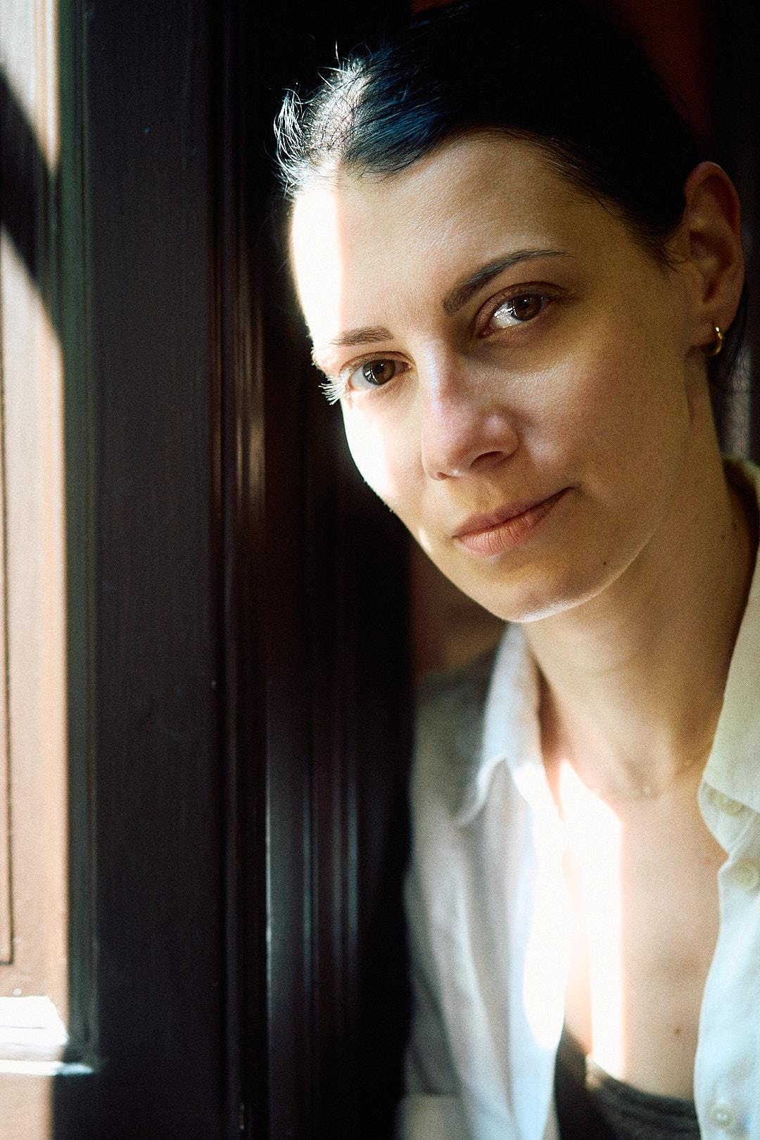 Portret kobiety w białej koszuli, oświetlonej promieniami słońca padającymi przez okno, z ciemnym framugą w tle. Subtelny makijaż, naturalny wyraz twarzy.
