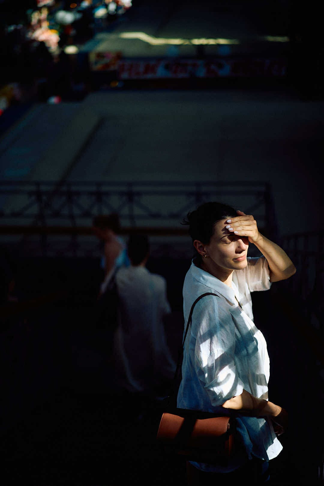 Portret kobiety w białej koszuli, osłaniającej oczy dłonią przed słońcem, z torbą na ramieniu. W tle zarys postaci i miejska sceneria w cieniu.