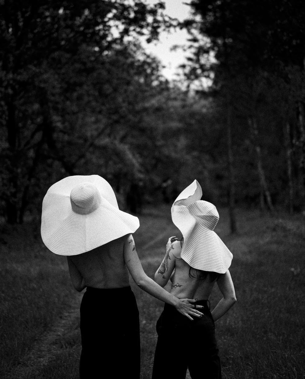 Dwie kobiety w kapeluszach o dużych rondach, widziane od tyłu, w czarno-białej sesji plenerowej na tle lasu. Jedna z tatuażem na ramieniu.