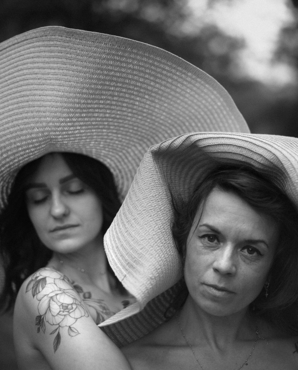 Portret dwóch kobiet w kapeluszach o szerokim rondzie, czarno-biała fotografia z widocznym tatuażem kwiatowym na ramieniu jednej z modelek.