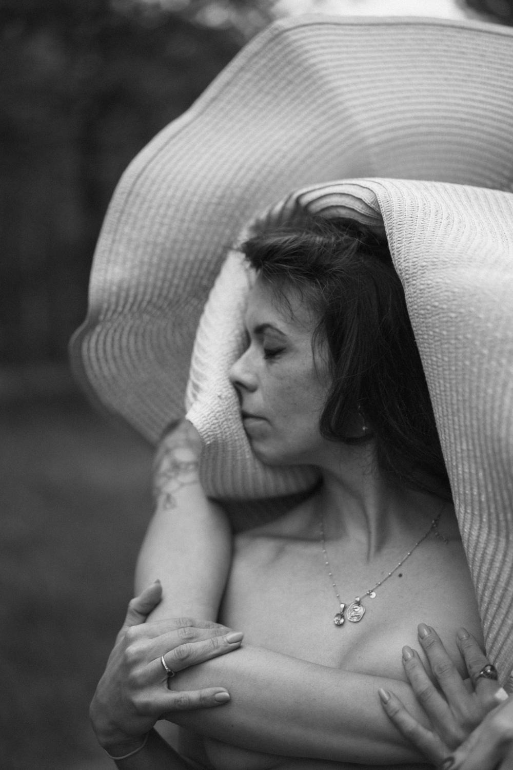 Czarno-białe, artystyczne ujęcie kobiety z zamkniętymi oczami, otulonej dużym, słomkowym kapeluszem. Widoczne delikatne detale, takie jak naszyjnik i pierścionek.