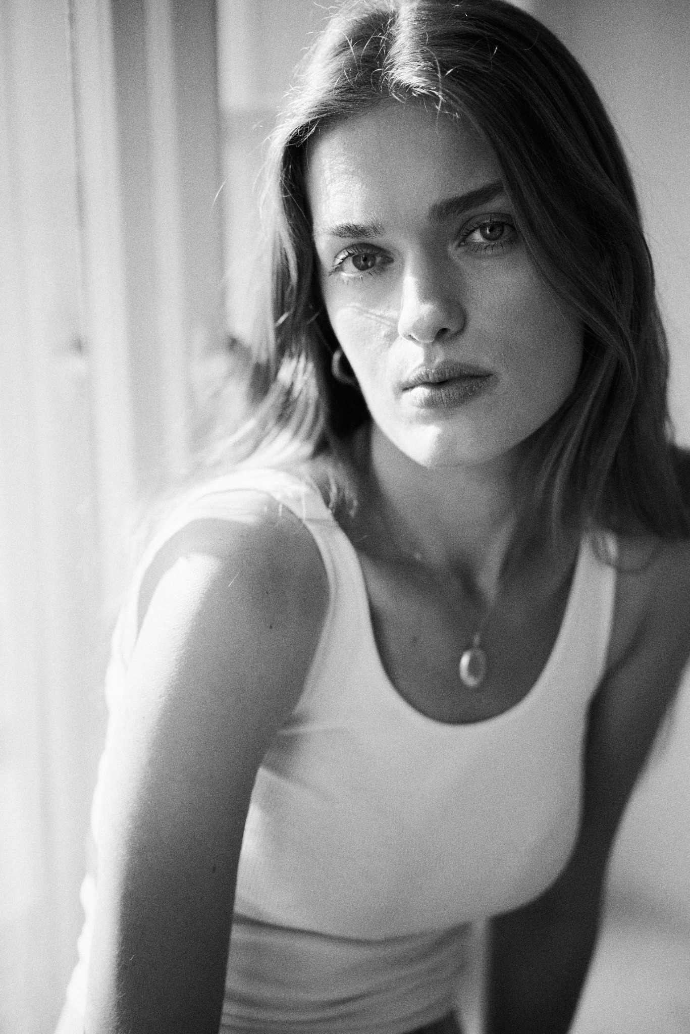 Czarno-biały portret kobiety w białym topie, z delikatnym naszyjnikiem, oświetlonej naturalnym światłem padającym z boku, tworzącym subtelne cienie na twarzy.