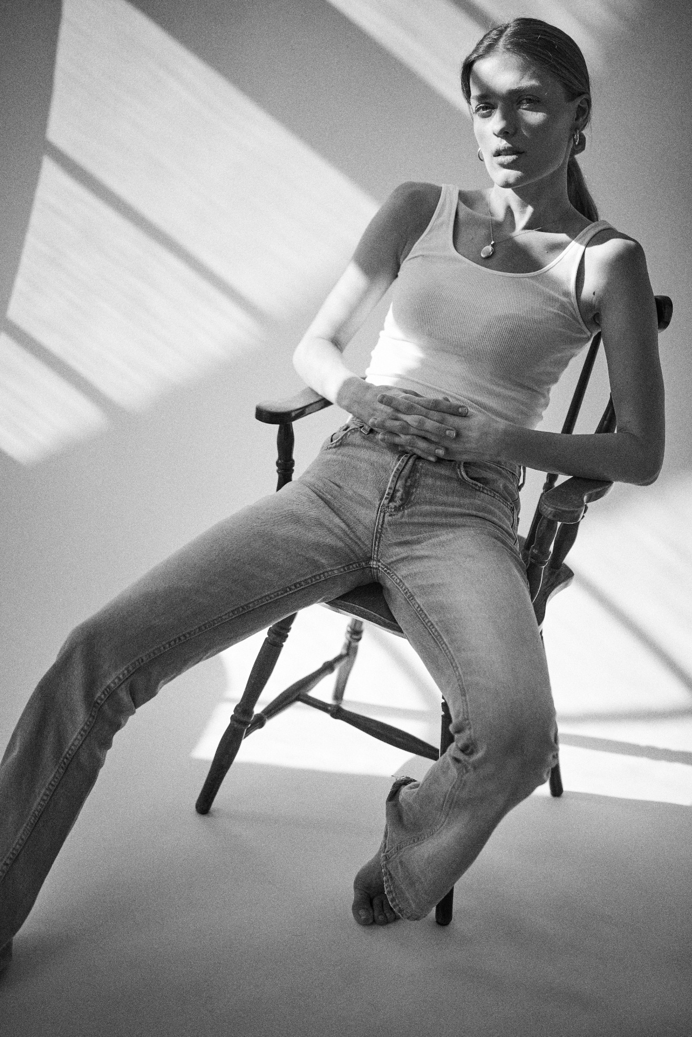 Czarno-białe zdjęcie kobiety siedzącej na krześle w dżinsach i podkoszulku, oświetlonej promieniami słońca wpadającymi przez okno, tworzącymi cienie na ścianie.