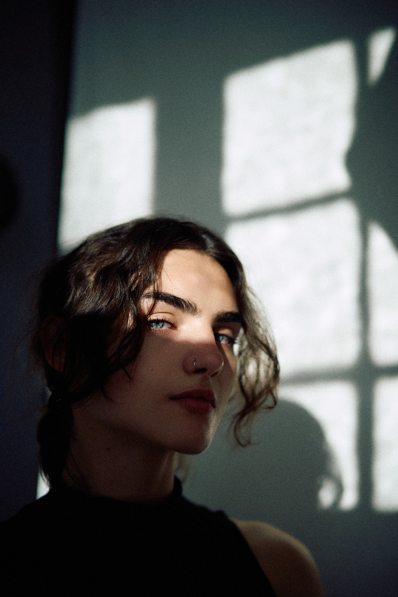 Portret kobiety o przenikliwym spojrzeniu, z kolczykiem w nosie, oświetlonej przez geometryczne cienie okna na ścianie. Ujęcie z bliska, podkreślające rysy twarzy.