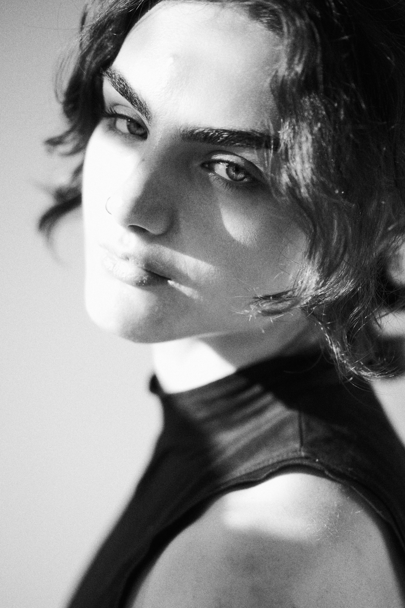 Czarno-biały portret kobiety z piercingiem w nosie, oświetlonej bocznym światłem, z delikatnym uśmiechem i kręconymi włosami, w bluzce z golfem.