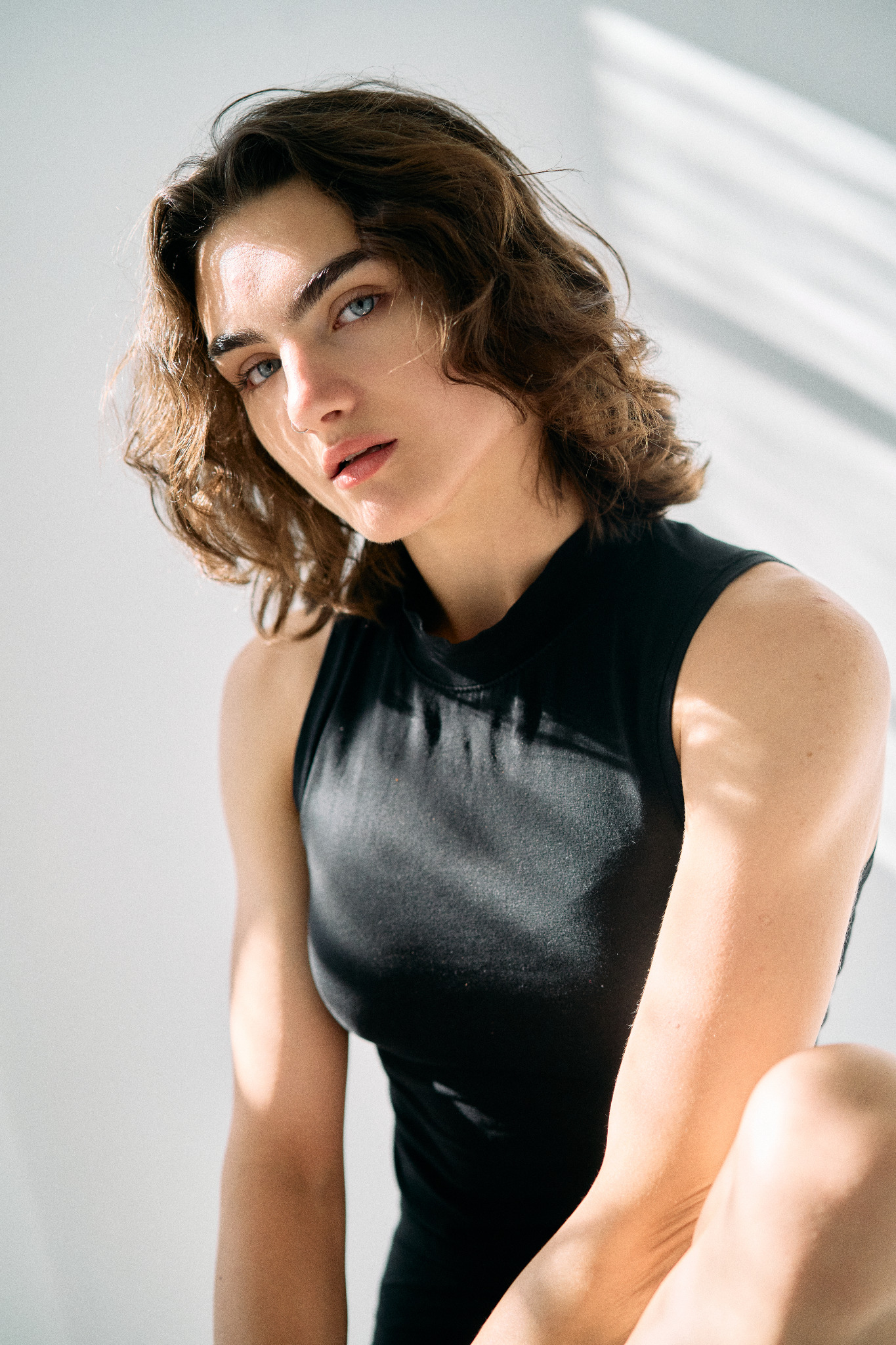 Portret kobiety o przenikliwym spojrzeniu, z krótkimi, kręconymi włosami i w czarnym topie, oświetlonej naturalnym światłem padającym przez okno.