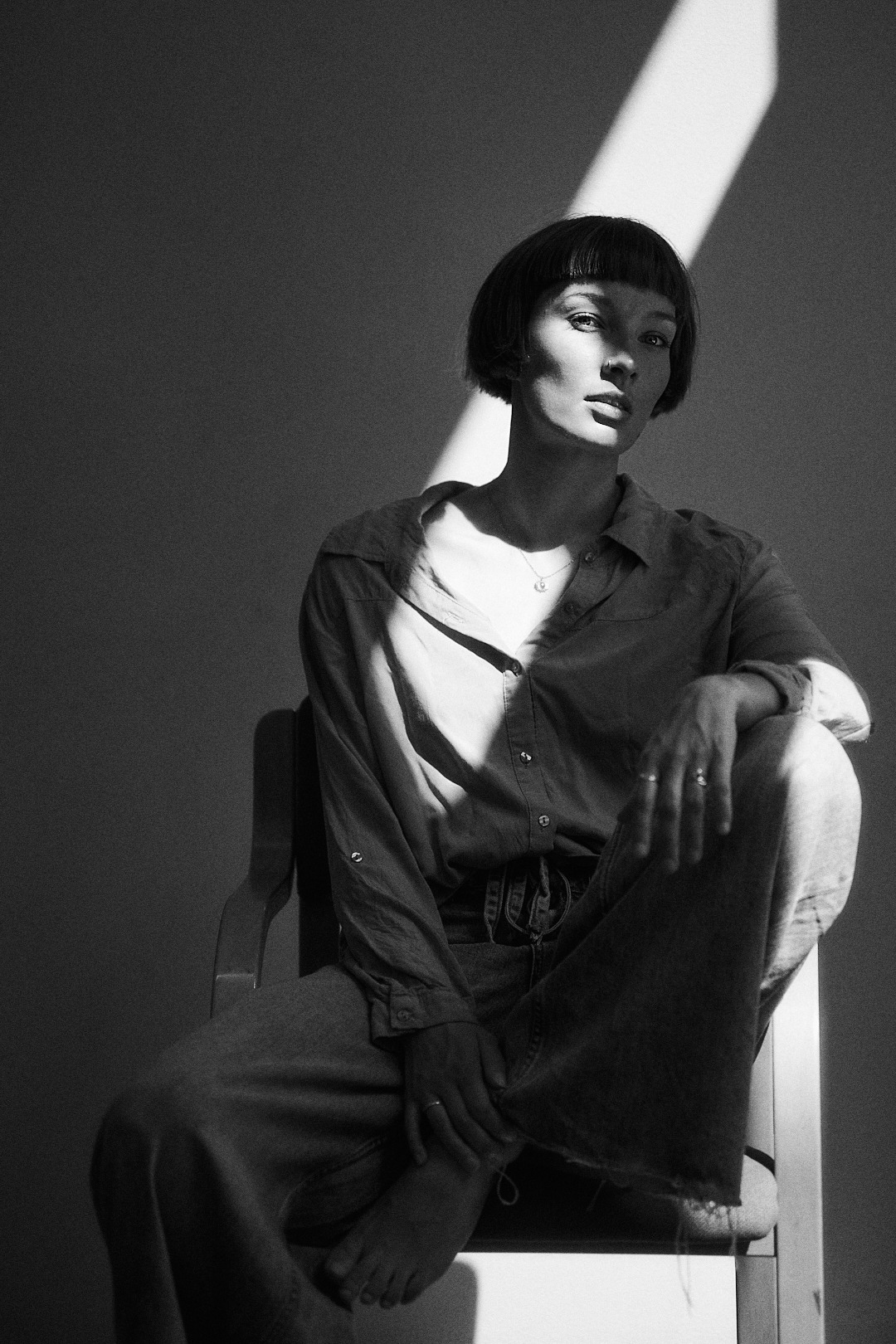 Czarno-biały portret kobiety z krótkimi włosami, siedzącej na krześle w silnym kontraście światła i cienia, z promieniem światła padającym na twarz.