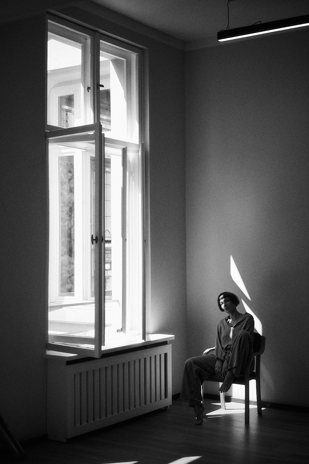 Monochromatyczne ujęcie kobiety siedzącej na krześle w pokoju z dużym oknem, na którą pada snop światła. Minimalistyczne wnętrze, wysoki kontrast, artystyczna fotografia.
