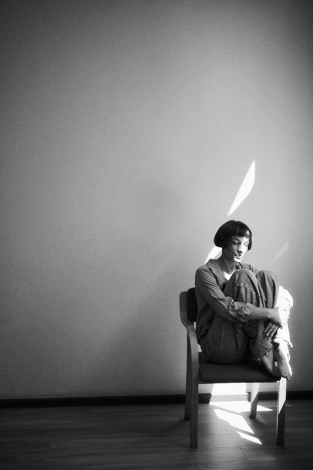 Monochromatyczne ujęcie kobiety z krótkimi włosami, siedzącej na krześle, obejmującej kolana, oświetlonej smugą światła na tle jasnej ściany.