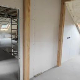 Montaż płyt gipsowo-kartonowych na drewnianej konstrukcji w poddaszu. Widoczne izolacje piankowe i elementy instalacji wentylacyjnej. Surowy stan wykończenia.