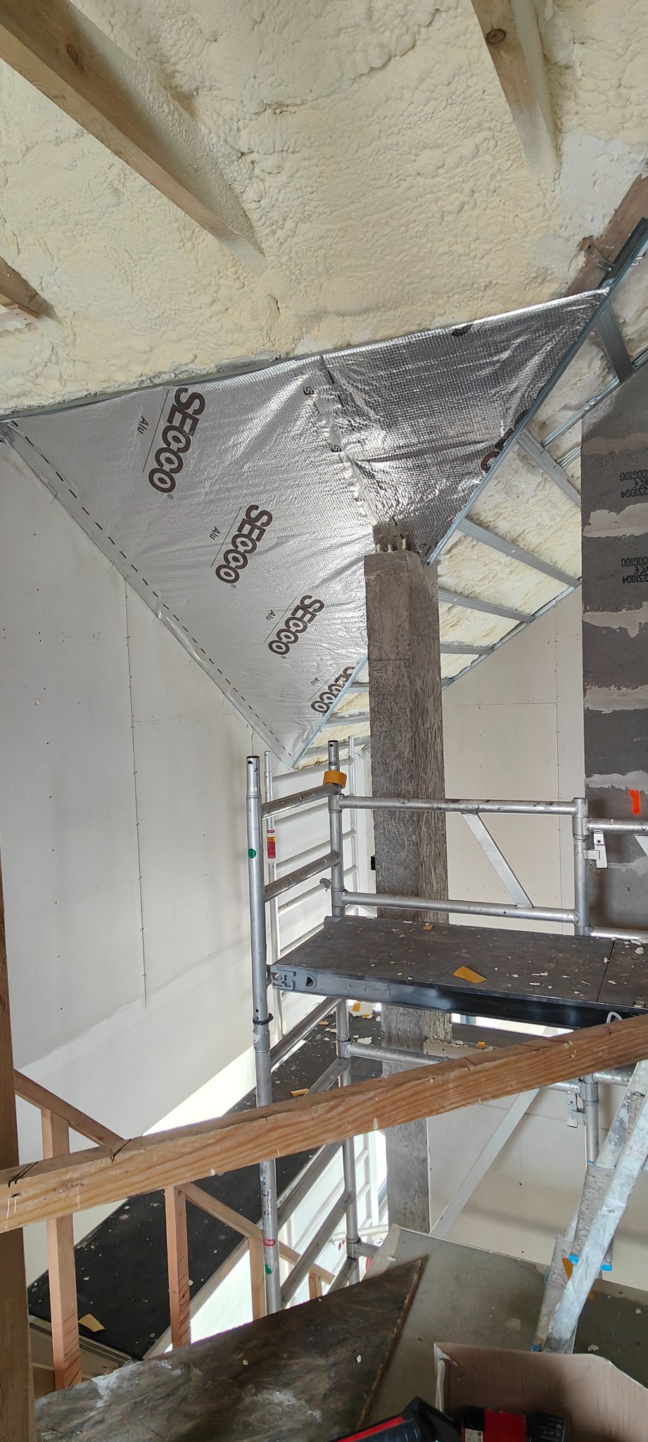 Ocieplenie poddasza pianą PUR i folią aluminiową SECOCO, widok z rusztowania. Elementy konstrukcyjne dachu i komin w surowym stanie. Prace w toku.