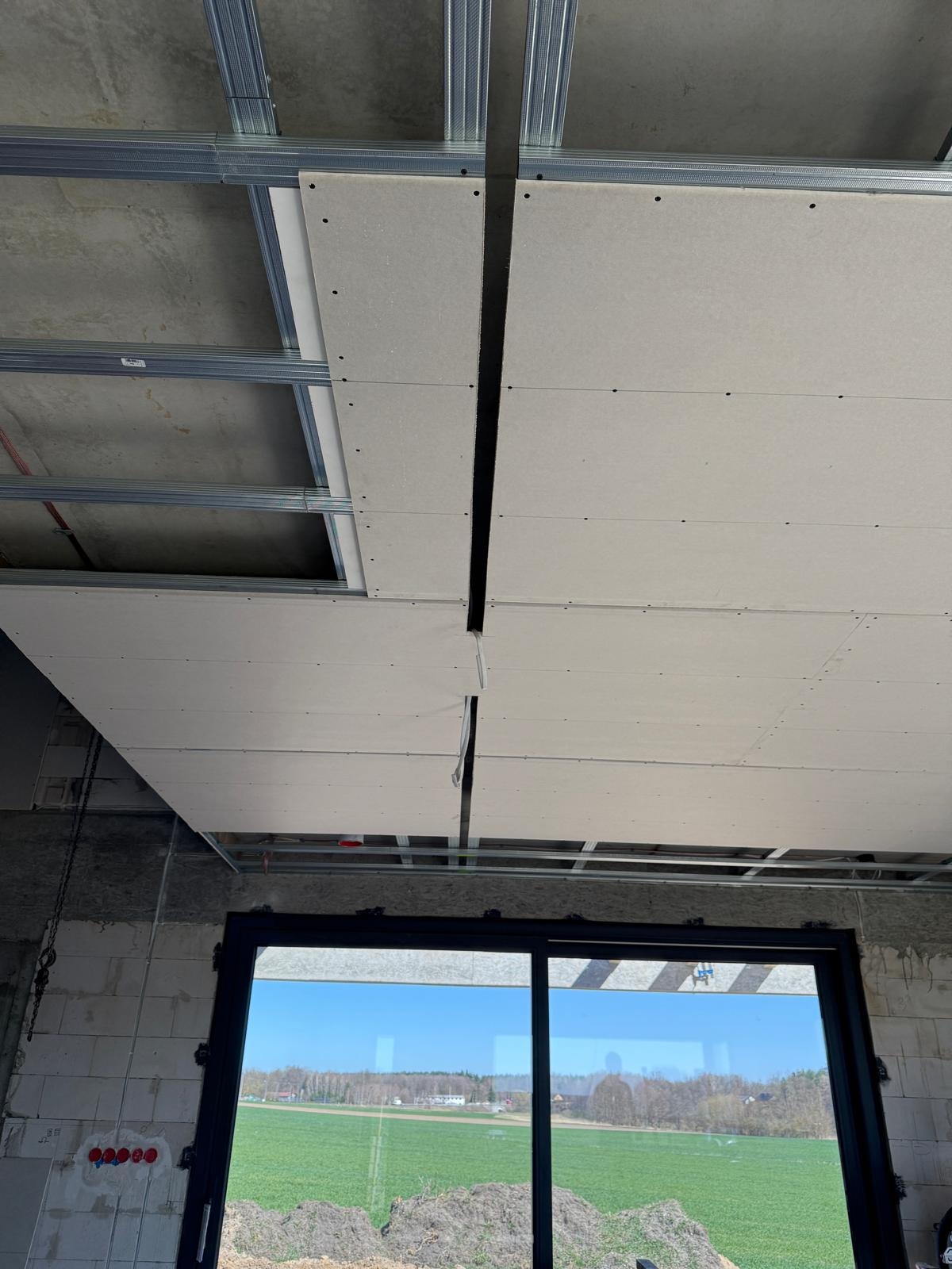 Montaż płyt karton-gips na metalowym ruszcie sufitowym w surowym wnętrzu z widokiem na krajobraz przez duże okno. Widoczne niedociągnięcia w łączeniach płyt.