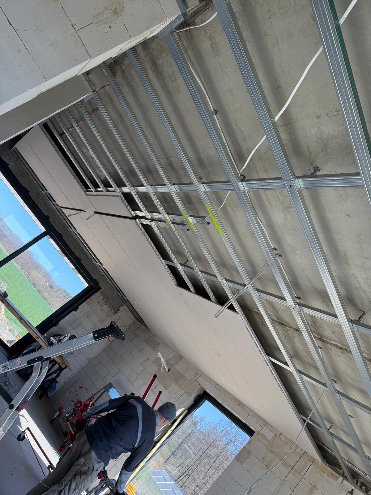 Montaż sufitu podwieszanego z płyt gipsowo-kartonowych na metalowej konstrukcji. Widoczne okna i częściowo zamontowane płyty. Fachowiec przy pracy.