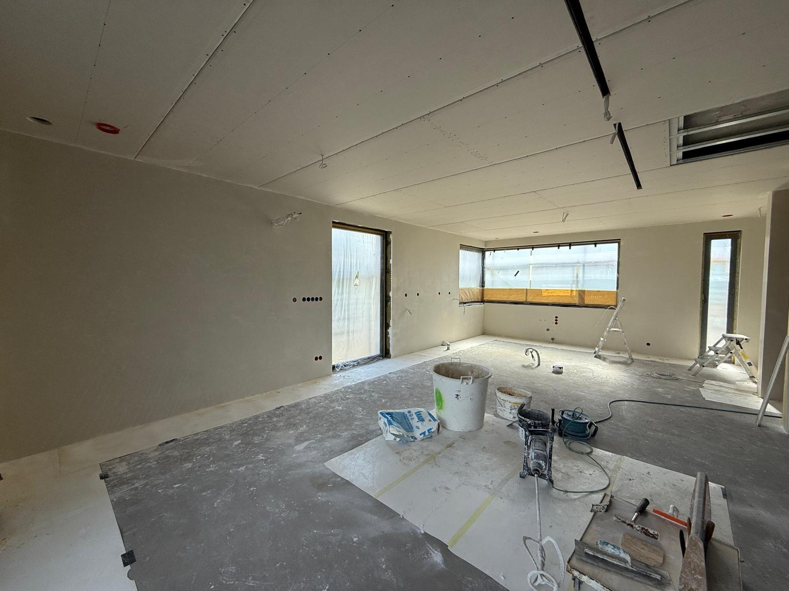 Wnętrze w trakcie szpachlowania i montażu płyt gipsowych. Widoczne narzędzia, drabiny, wiaderka z zaprawą, oraz okna zabezpieczone folią malarską.