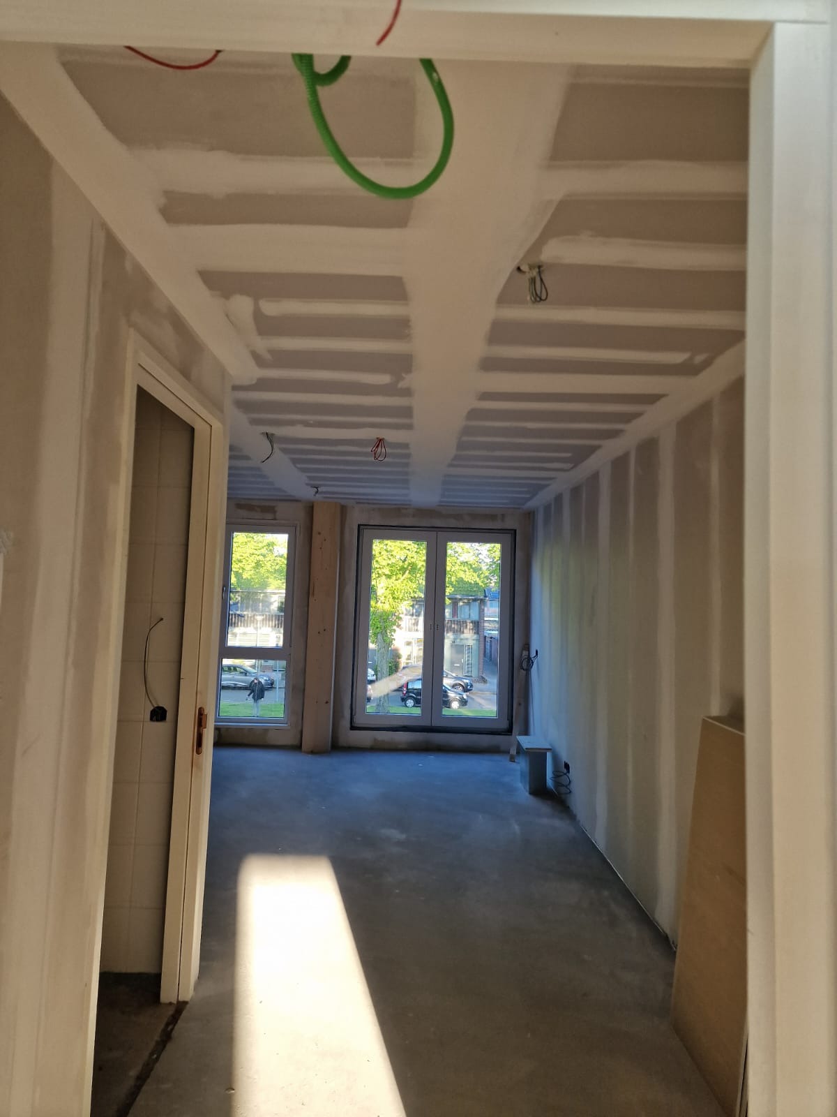 Wnętrze w trakcie prac: płyty gipsowo-kartonowe na ścianach i suficie, widoczne instalacje elektryczne. Dwa okna balkonowe z widokiem na zieleń. Stan surowy przed wykończeniem.