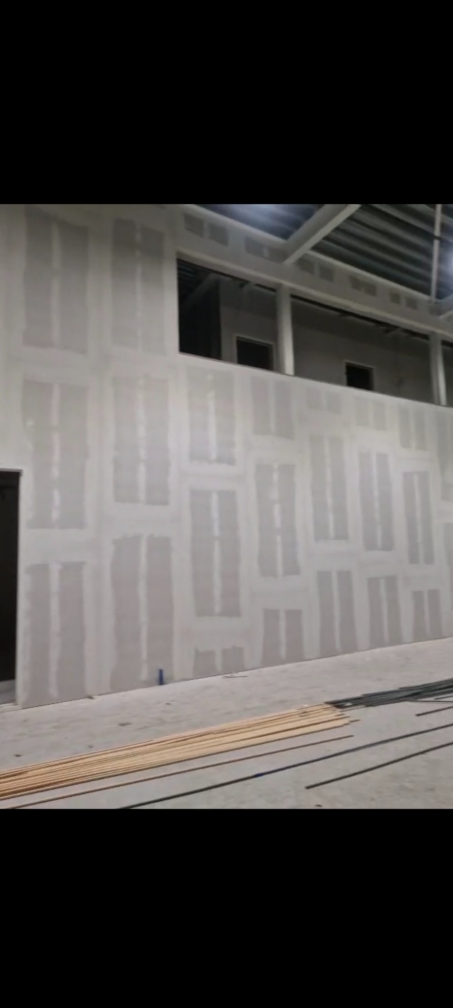 Wysoka ściana z płyt gipsowo-kartonowych, częściowo wykończona szpachlą, w dużym pomieszczeniu z widoczną konstrukcją stalową dachu i materiałami budowlanymi na podłodze.