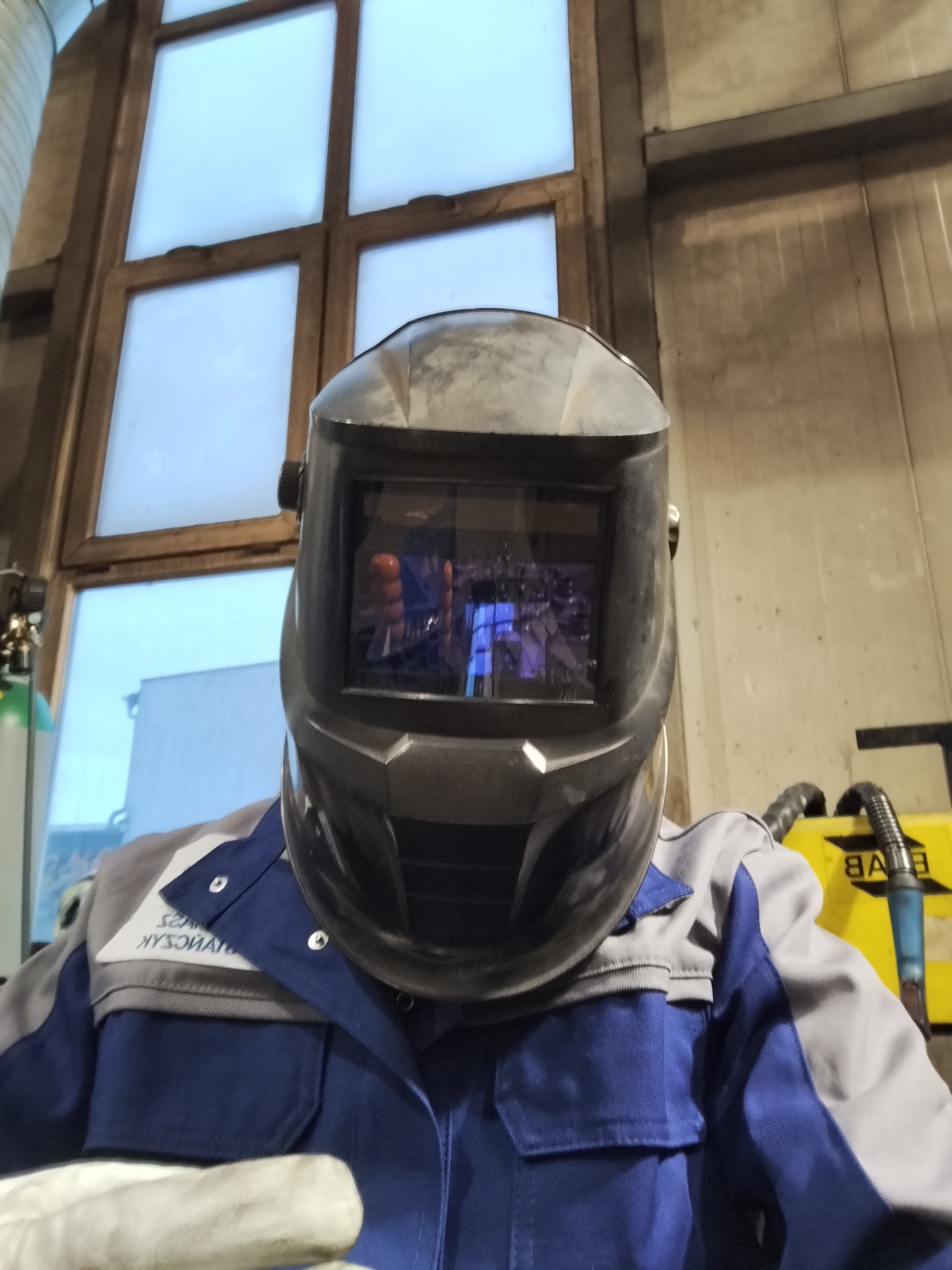 Spawacz w masce ochronnej, widoczny fragment okna i sprzęt spawalniczy w tle. Ujęcie z dołu, podkreślające ochronę podczas pracy.