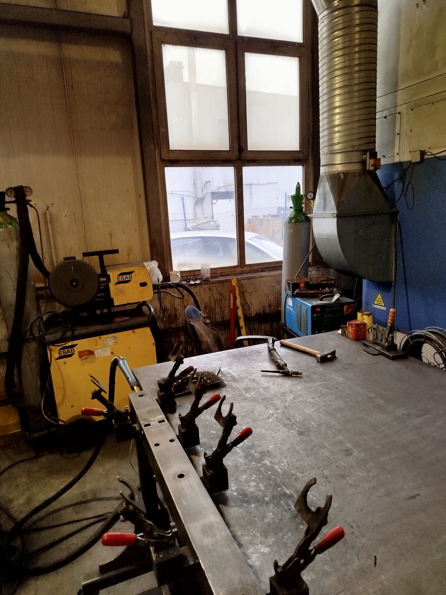 Warsztat ślusarski: metalowy stół z narzędziami, spawarka ESAB, butla z gazem, okno z widokiem na zewnątrz. Przygotowania do pracy.