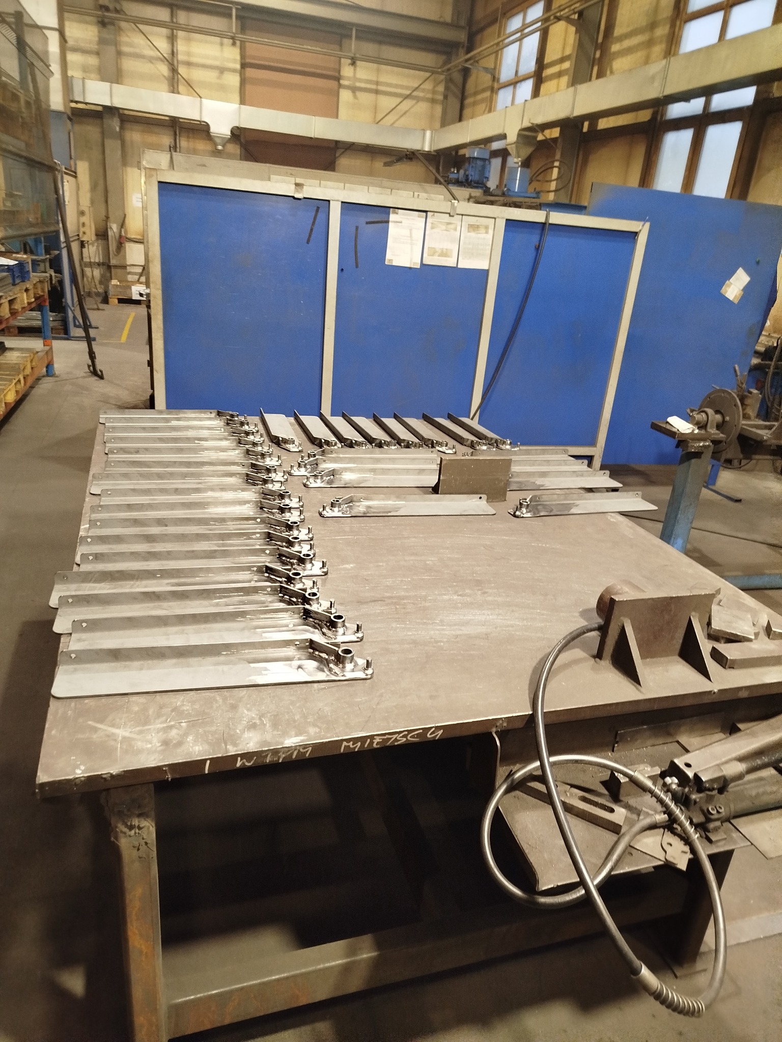 Metalowe elementy z uchwytami ułożone rzędami na stole warsztatowym w hali produkcyjnej, przygotowane do dalszej obróbki lub montażu.