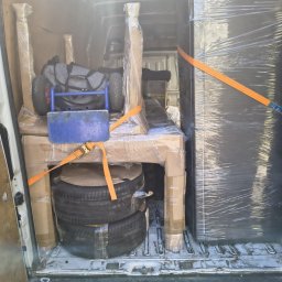 Usługi Transportowe i Przewóz Paczek Tomasz Grabarz - Wnętrze busa załadowane meblami, oponami i wózkiem transportowym, zabezpieczonymi folią i pasami. Widok z tyłu pojazdu, gotowy do transportu.