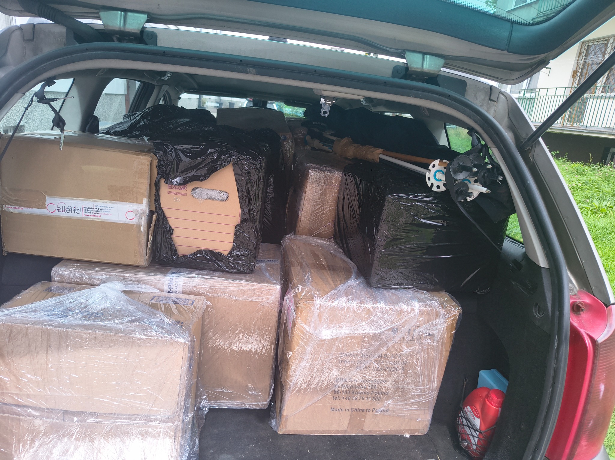 Kartonowe pudła, niektóre owinięte folią i czarną folią, wypełniają bagażnik samochodu typu kombi podczas transportu. Widoczne elementy wyposażenia wnętrza pojazdu.