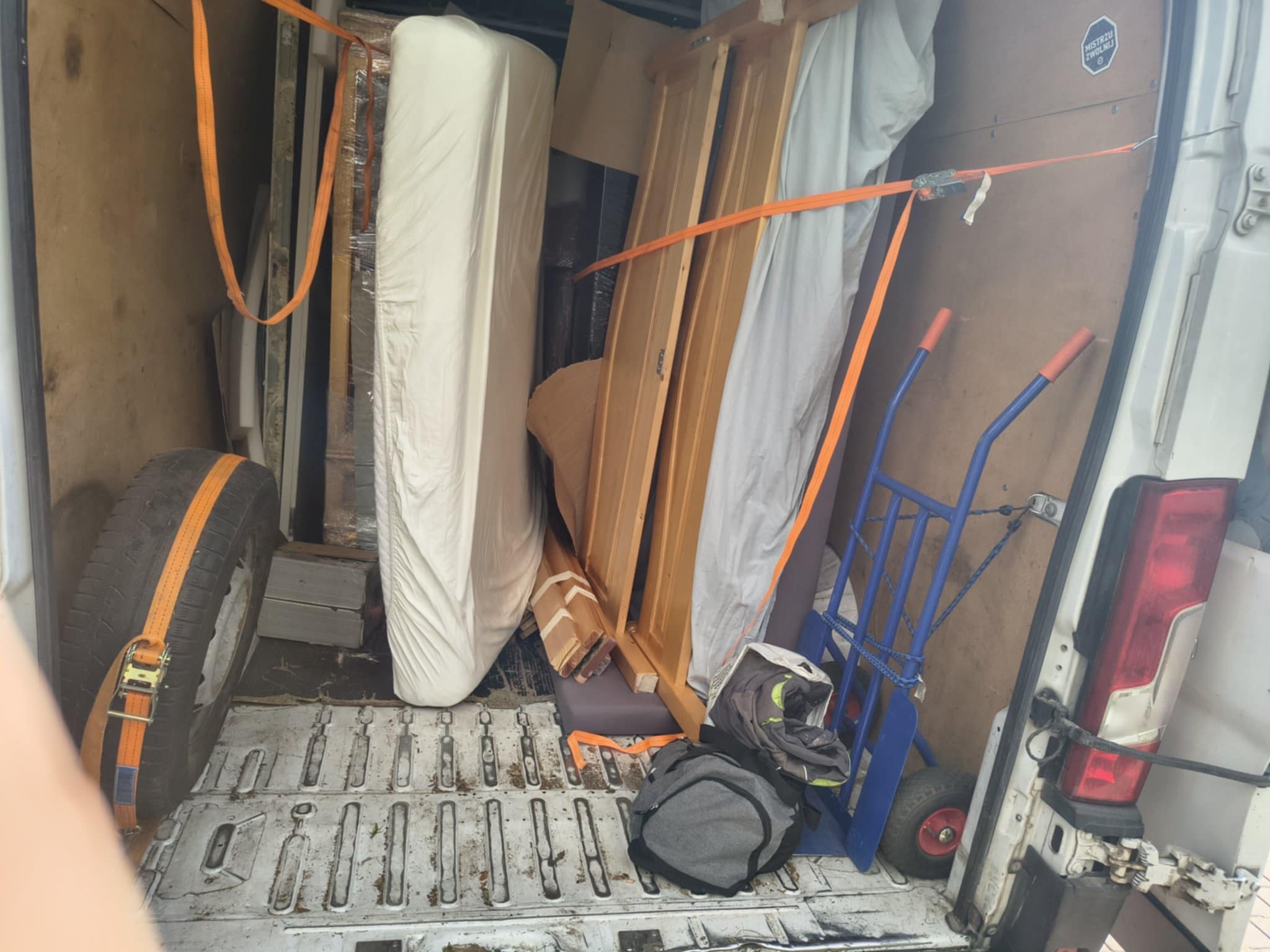 Wnętrze busa załadowane meblami i materacem, zabezpieczonymi pomarańczowymi pasami transportowymi. Widoczny wózek transportowy i koło zapasowe.