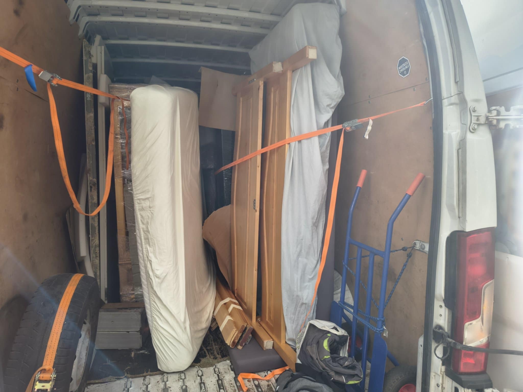 Wnętrze busa załadowane meblami i sprzętami, zabezpieczone pomarańczowymi pasami transportowymi. Widoczny wózek transportowy i opona.