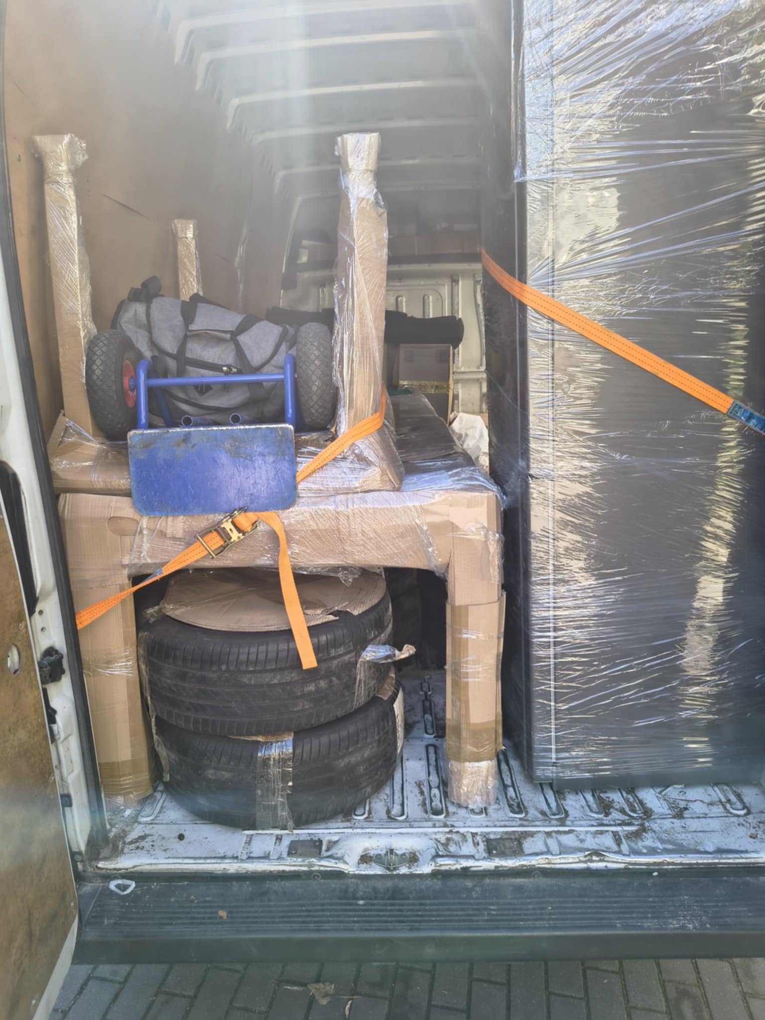 Wnętrze busa załadowane meblami, oponami i wózkiem transportowym, zabezpieczonymi folią i pasami. Widok z tyłu pojazdu, gotowy do transportu.