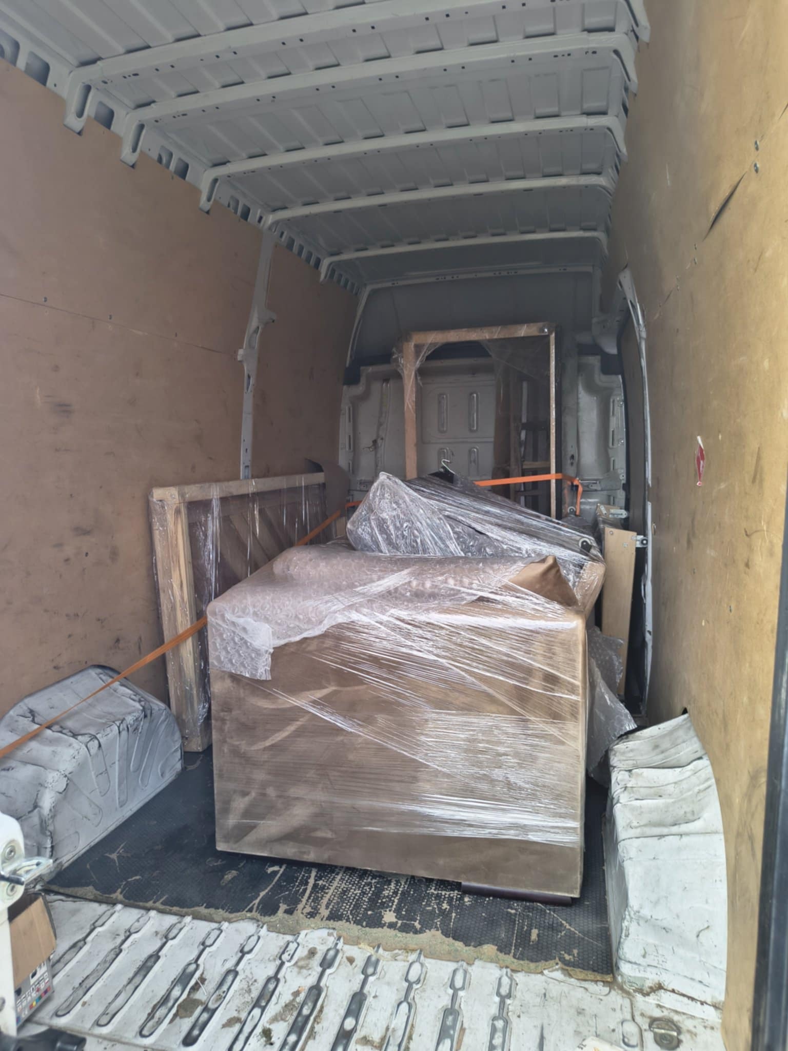 Wnętrze furgonetki wypełnionej meblami i kartonami zabezpieczonymi folią stretch i pasami transportowymi. Widok z tyłu pojazdu, jasne oświetlenie.