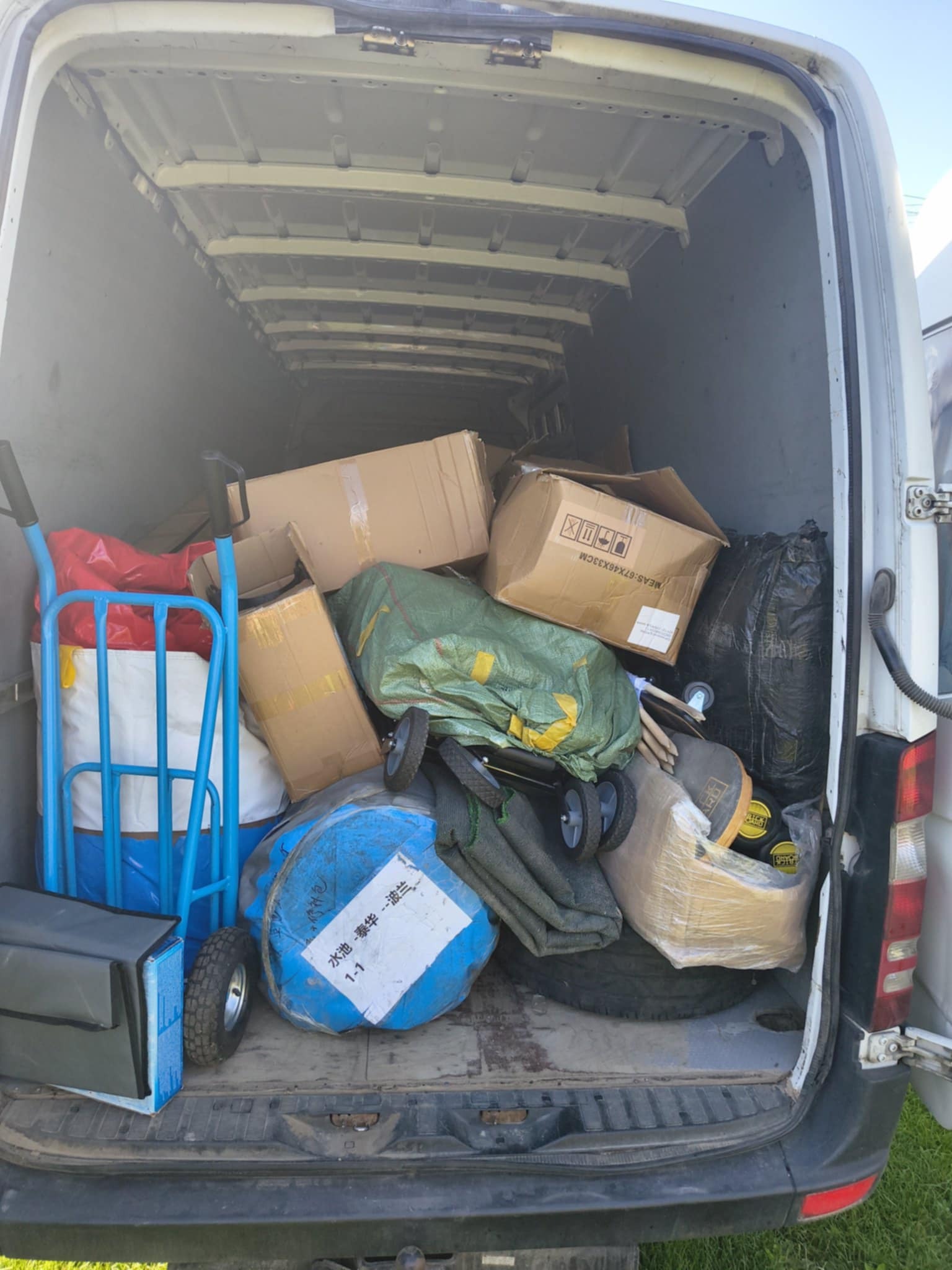 Wnętrze białego busa wypełnione kartonami, workami i wózkiem transportowym. Widoczne różne przedmioty przygotowane do przewozu. Ujęcie z tyłu pojazdu.