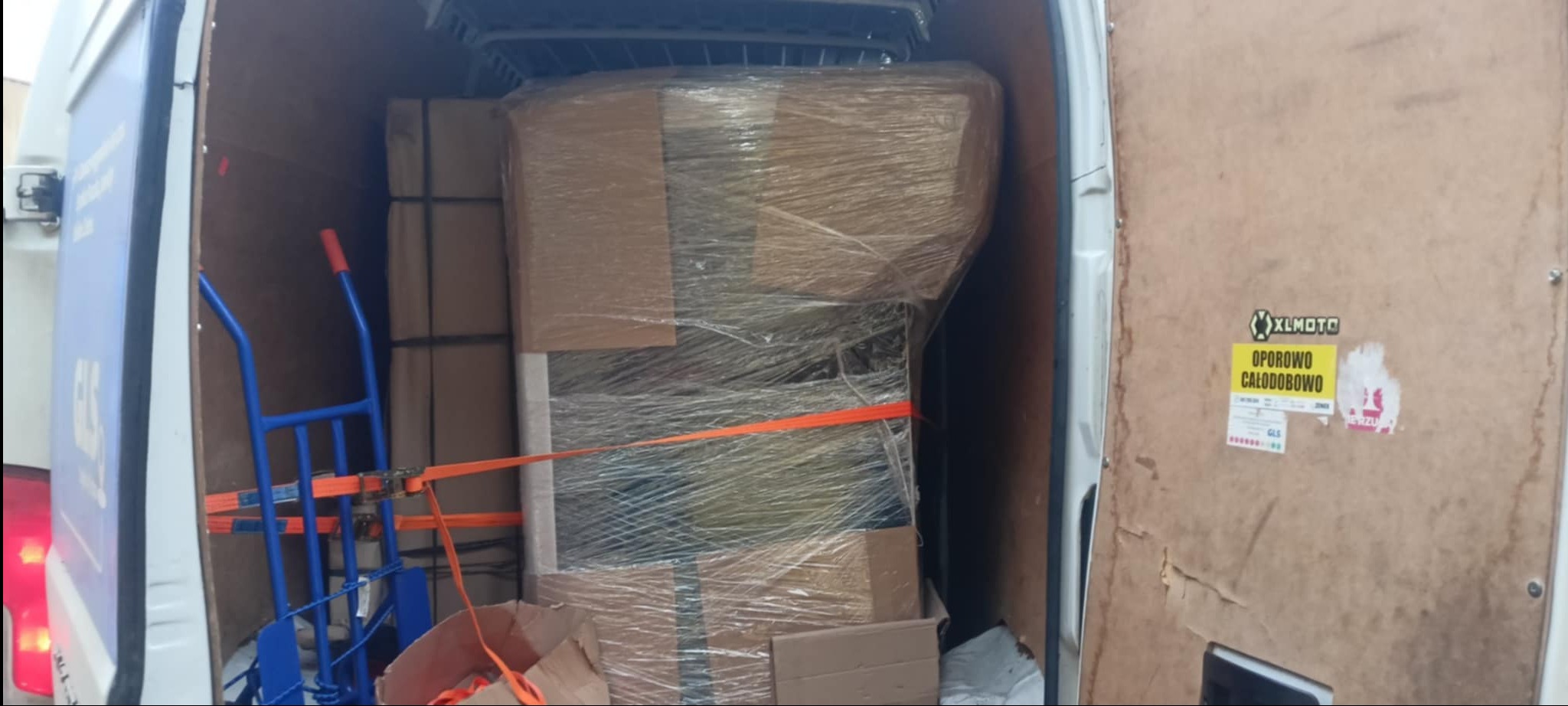 Wnętrze busa z załadowanymi, zabezpieczonymi kartonami i wózkiem transportowym. Widoczne pasy mocujące. Naklejki na drzwiach busa.