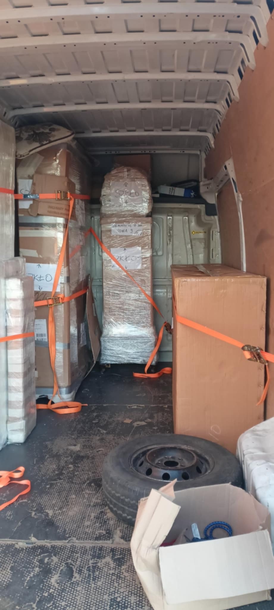 Wnętrze busa wypełnione kartonami i meblami zabezpieczonymi pasami transportowymi. Widoczne oznaczenia 'szkło' na paczkach. Koło zapasowe na podłodze.