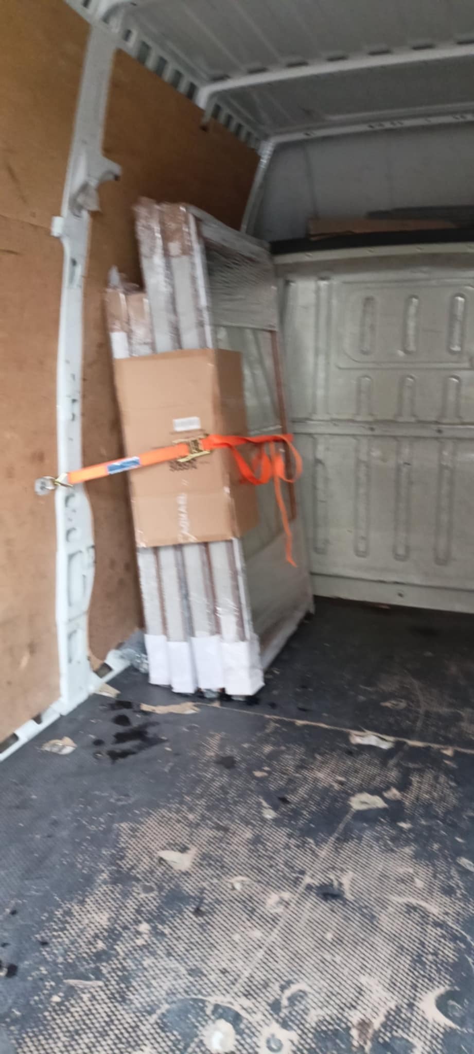 Wnętrze busa z zabezpieczonym ładunkiem: okna w folii i karton, spięte pomarańczowym pasem transportowym. Widoczna podłoga i ściany pojazdu.