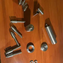 Zestaw precyzyjnie wykonanych, metalowych elementów o różnych kształtach, leżących na drewnianej powierzchni. Widoczne gwinty, otwory i gładkie wykończenie.