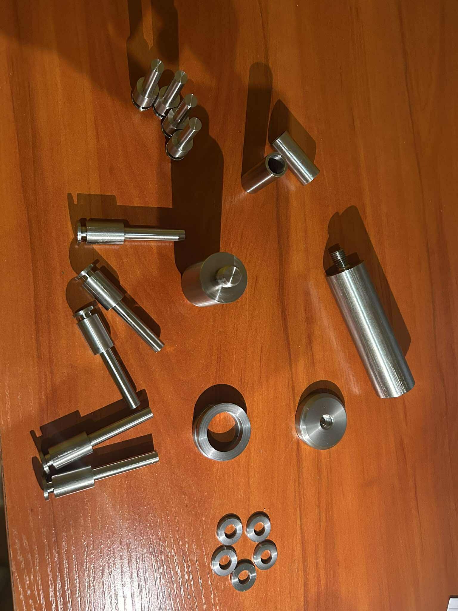 Zestaw precyzyjnie wykonanych, metalowych elementów o różnych kształtach, leżących na drewnianej powierzchni. Widoczne gwinty, otwory i gładkie wykończenie.