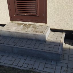 Usługi budowlane Maciej Markiewicz - Ułożone schody wejściowe z kostki brukowej w odcieniach szarości, narzędzia pracy obok, na zewnątrz budynku z beżową elewacją i brązową kratką wentylacyjną.