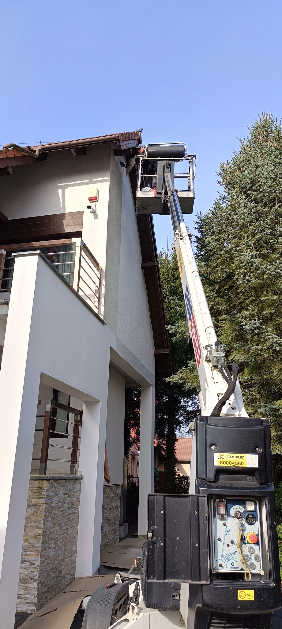 Pracownik na podnośniku koszowym maluje elewację domu jednorodzinnego. Widok na fragment budynku, drzewo i błękitne niebo.