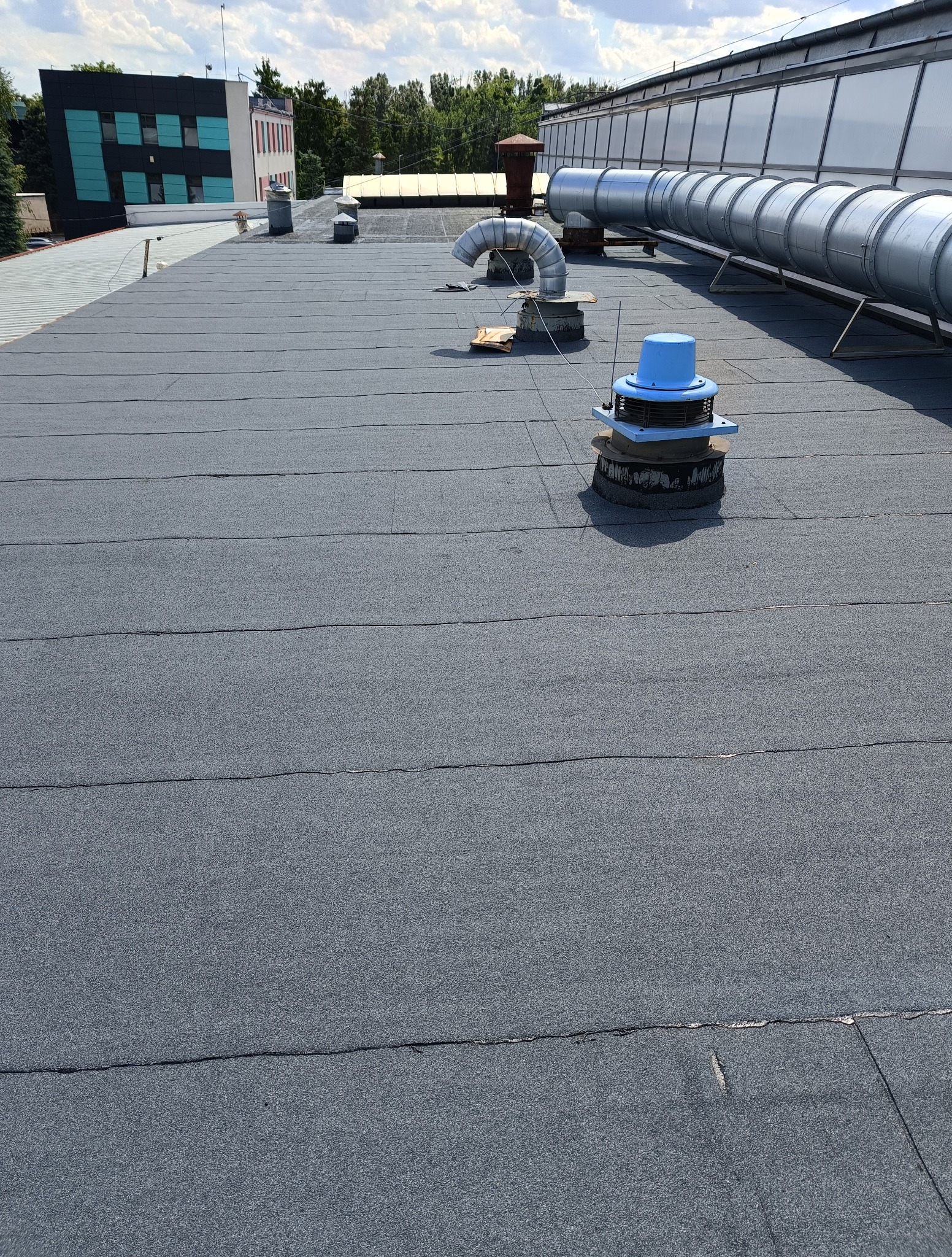 Szczegółowe ujęcie płaskiego dachu pokrytego papą termozgrzewalną z elementami wentylacji w Elblągu. Widoczne łączenia i faktura materiału, budynki w tle.