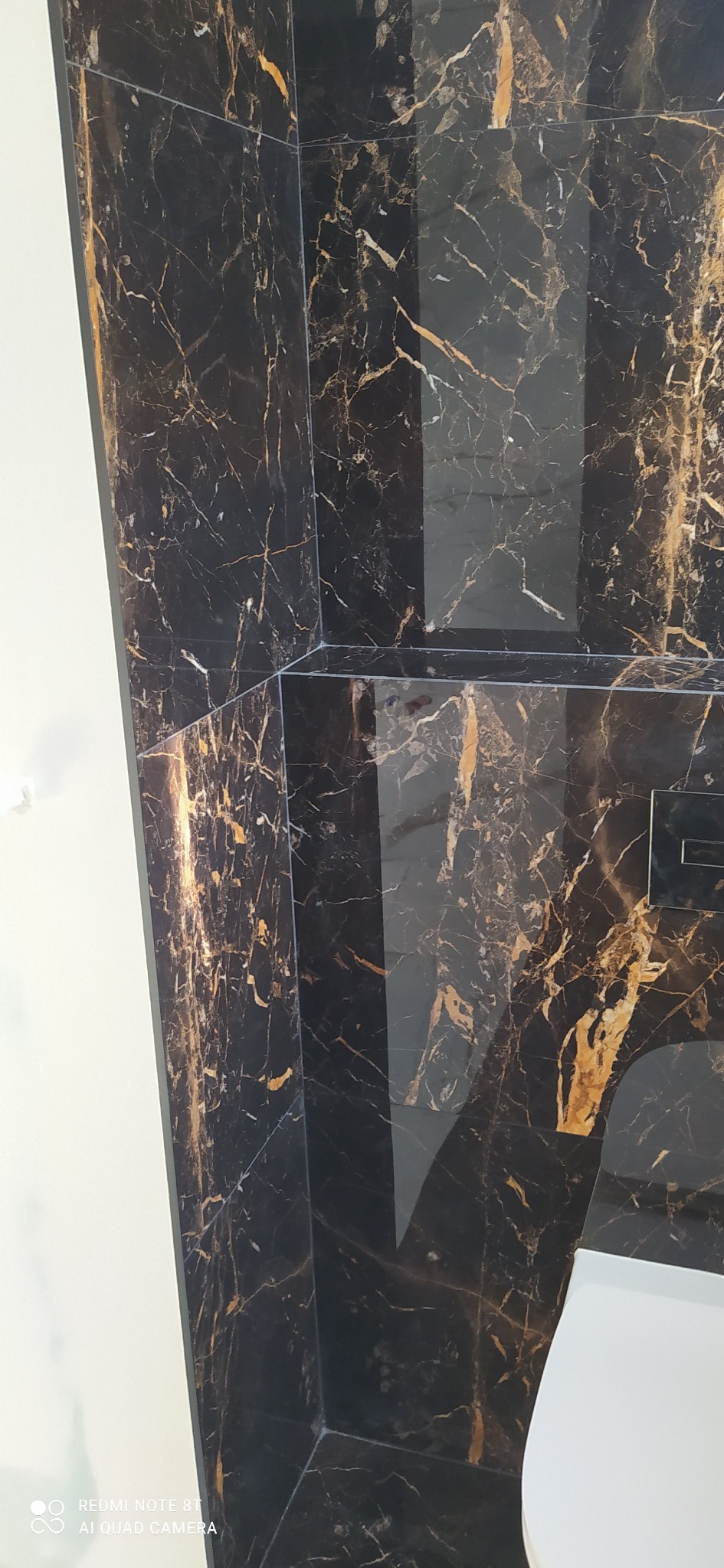 Czarne płytki imitujące marmur ze złotymi żyłami, ułożone na ścianie w łazience, widok z dołu, fragment toalety z widocznym przyciskiem spłukiwania.