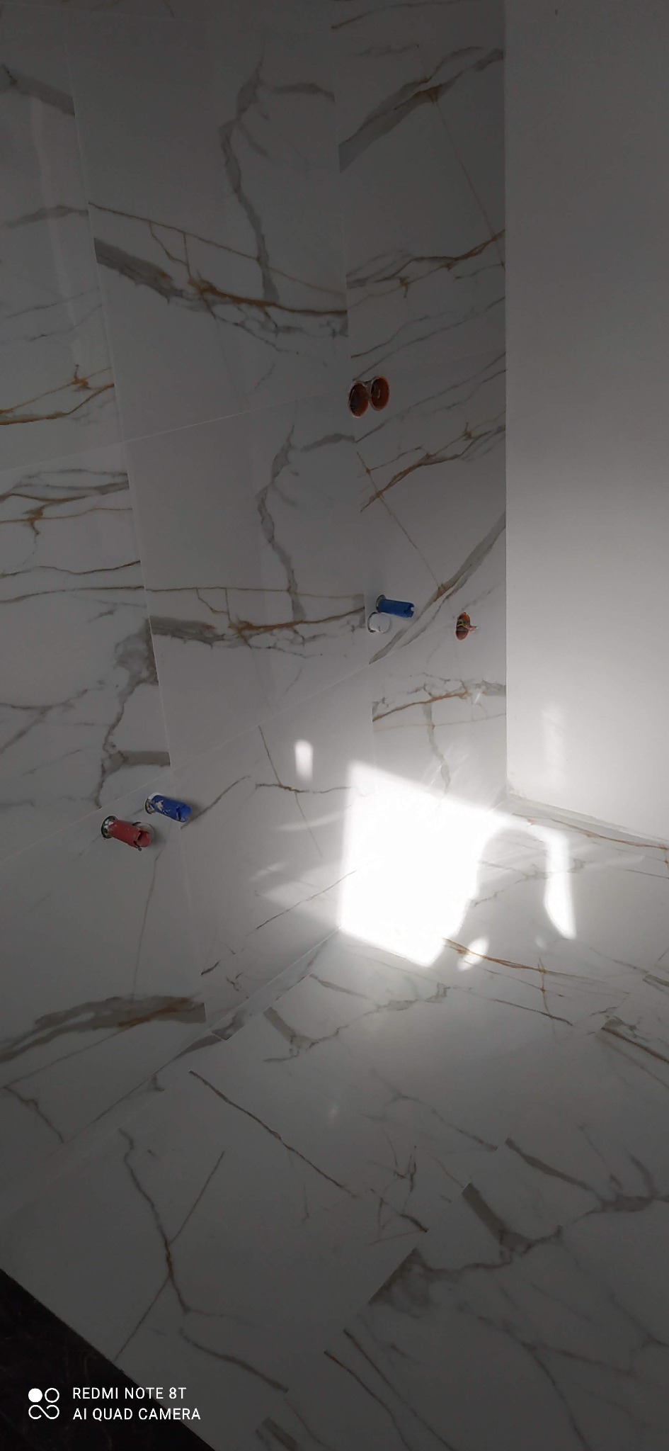 Białe płytki imitujące marmur z złotymi żyłami, ułożone na ścianie i podłodze łazienki, widoczne przyłącza wodne.