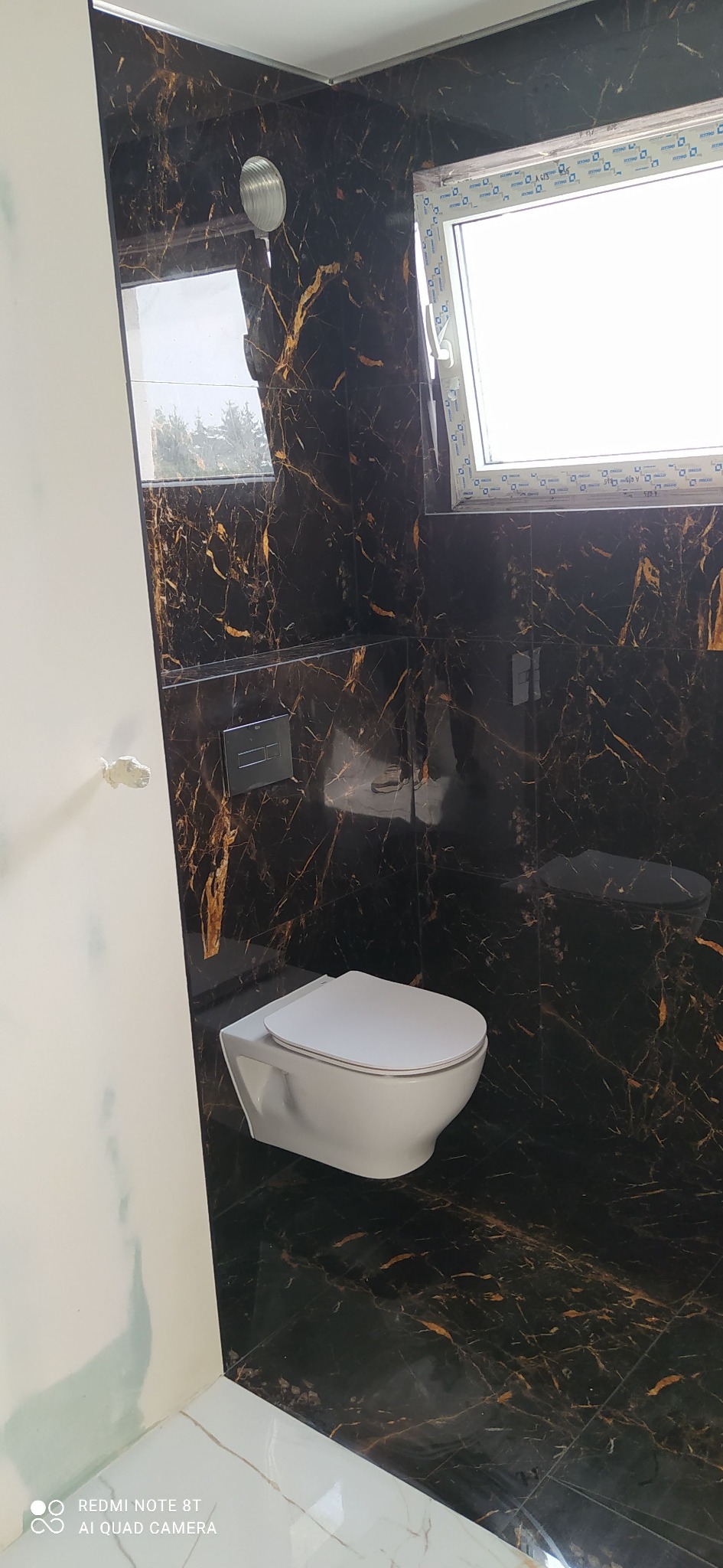 Wykończona łazienka z czarnymi płytkami imitującymi marmur ze złotymi żyłami, wisząca toaleta i okno. Nowoczesny design z akcentem luksusu.