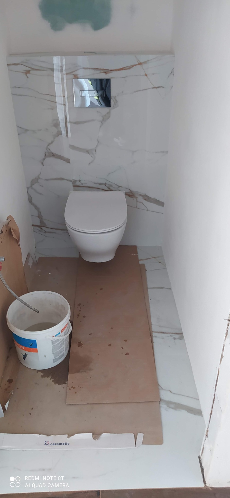 Biała toaleta podtynkowa w trakcie montażu w małej łazience wyłożonej płytkami imitującymi marmur. Widoczne wiadro i płyty zabezpieczające podłogę.