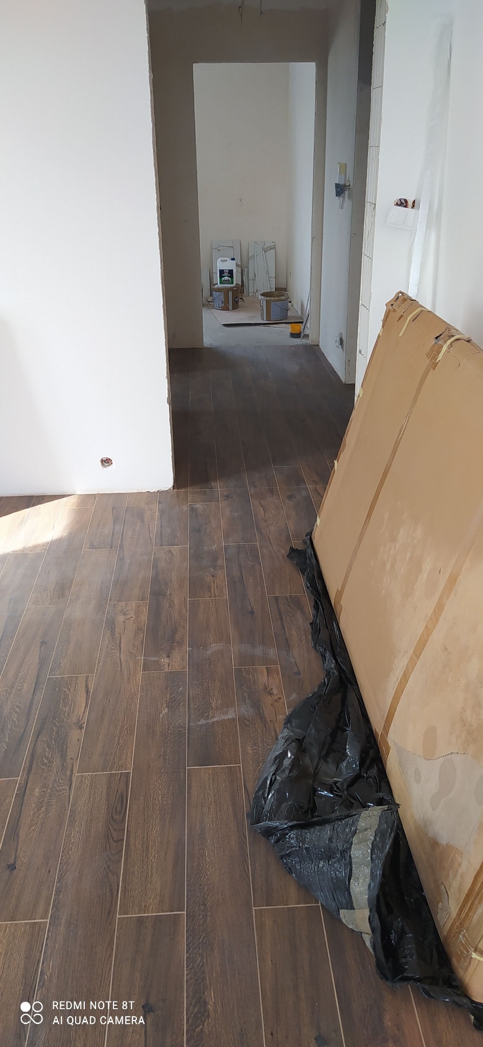 Ułożona podłoga z paneli imitujących drewno w jasnym wnętrzu. Widoczne kartony i czarna folia zabezpieczająca. W tle pomieszczenia w trakcie remontu.