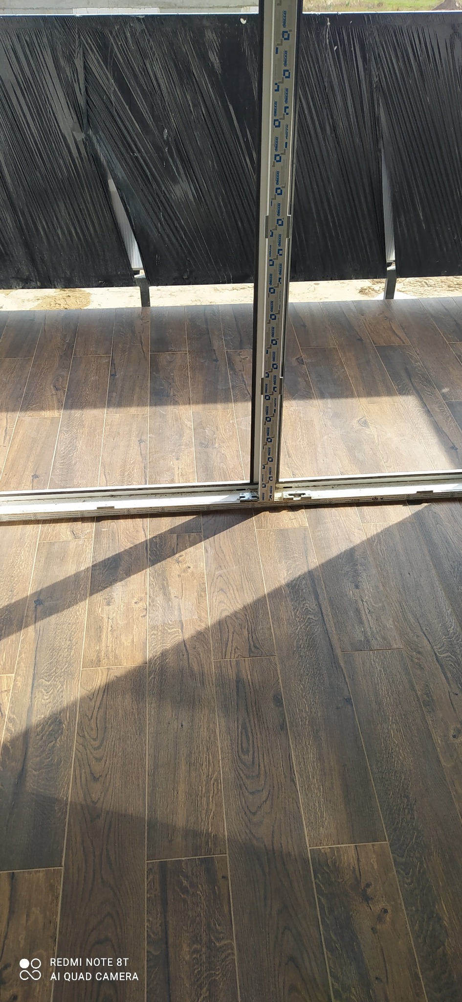 Ułożone panele podłogowe w odcieniach brązu, widoczne w świetle słonecznym. Częściowo zasłonięte folią ochronną i ramą okienną.