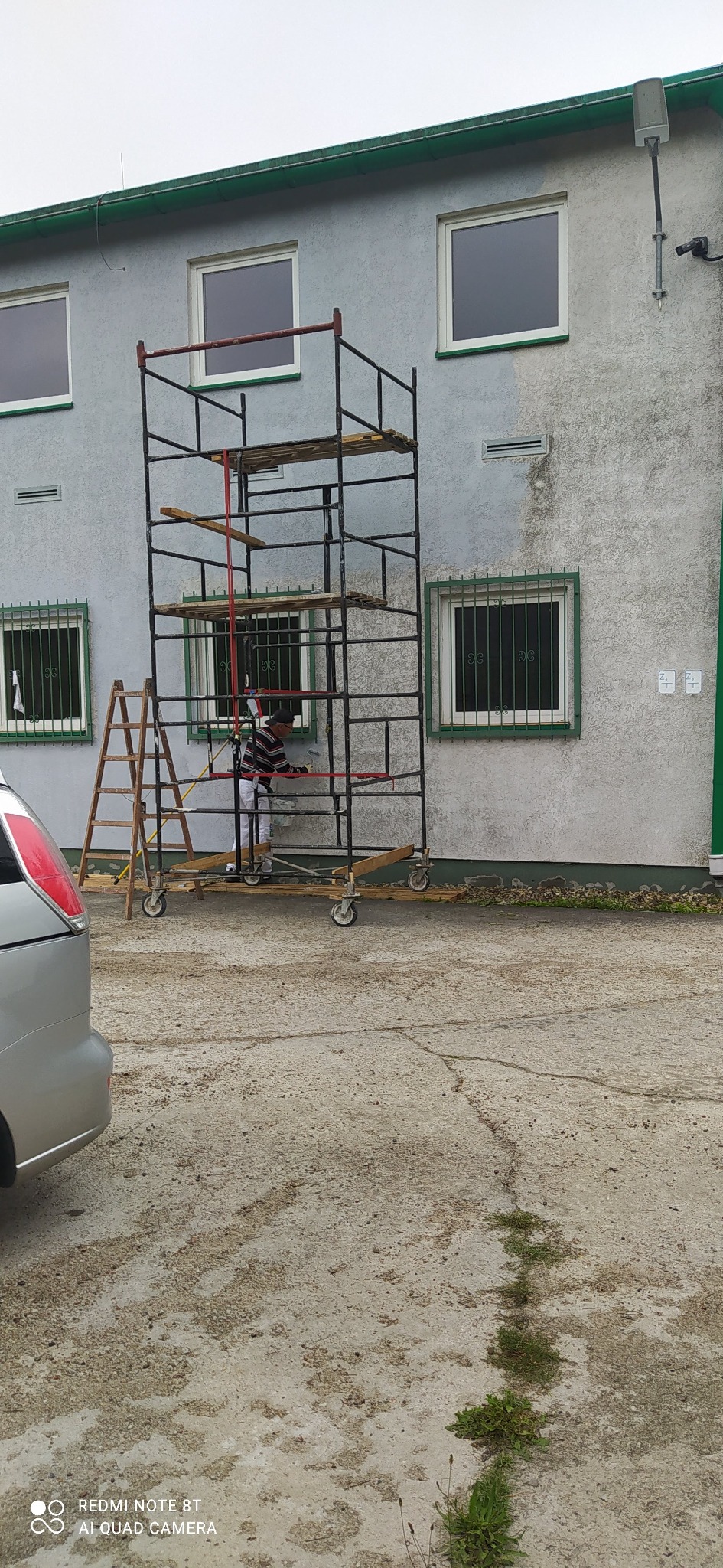 Pracownik na rusztowaniu maluje elewację budynku. Widoczna drabina, fragment samochodu i szara, chropowata ściana. Ujęcie z dołu, przy pochmurnej pogodzie.
